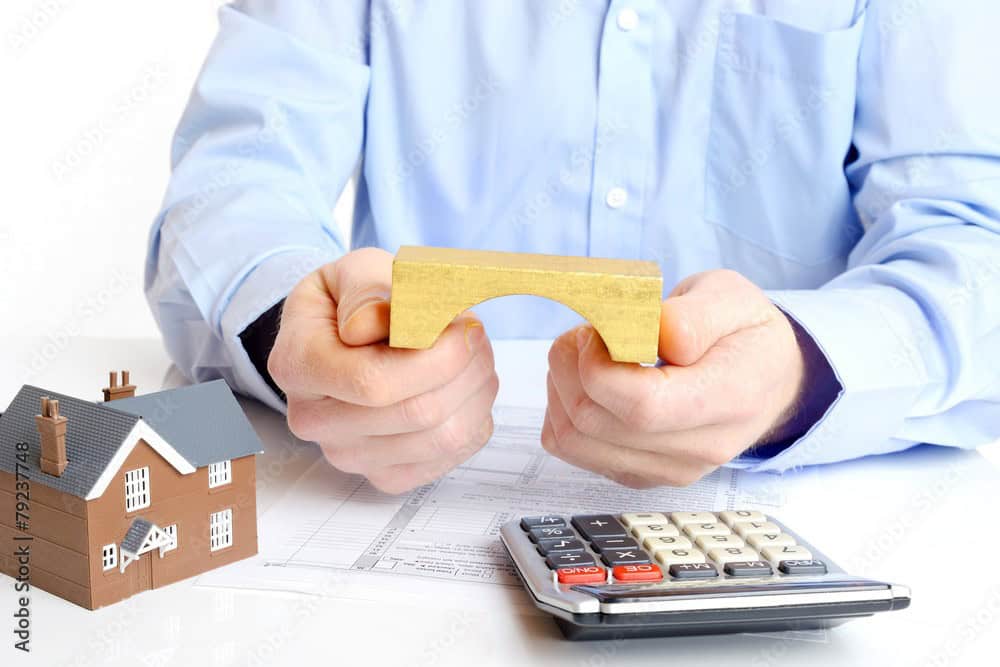 Hands holding a small wooden bridge between a model house and a calculator