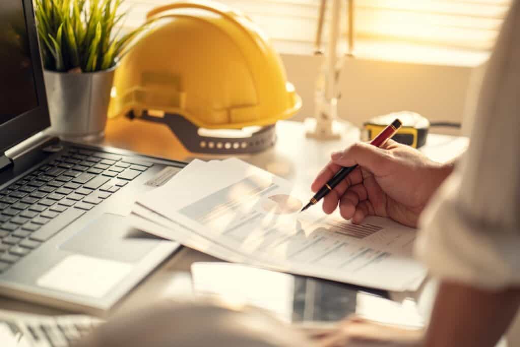 Person reviewing plans at a desk beside a yellow hard hat and a laptop
