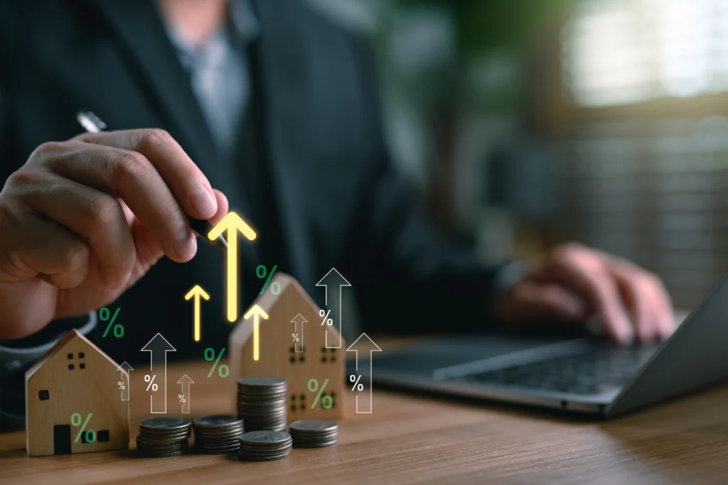Hand stacking coins beside model houses with upward arrows
