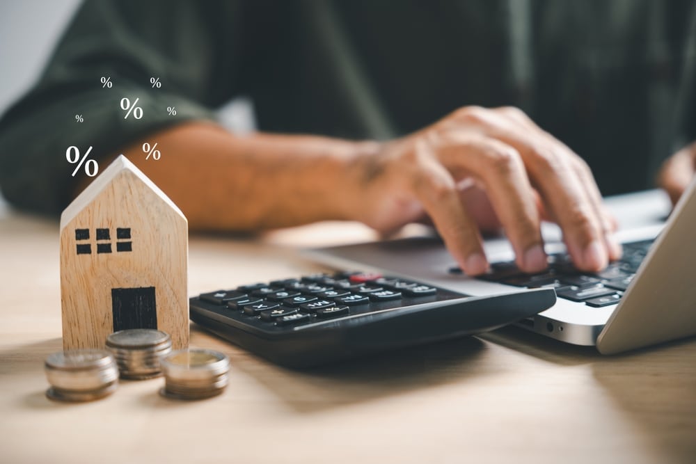Hand on a calculator beside a wooden model house and coins