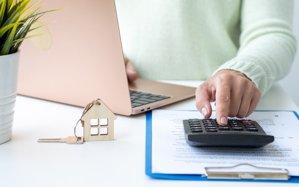 Hand on a calculator beside a house key ring and a laptop