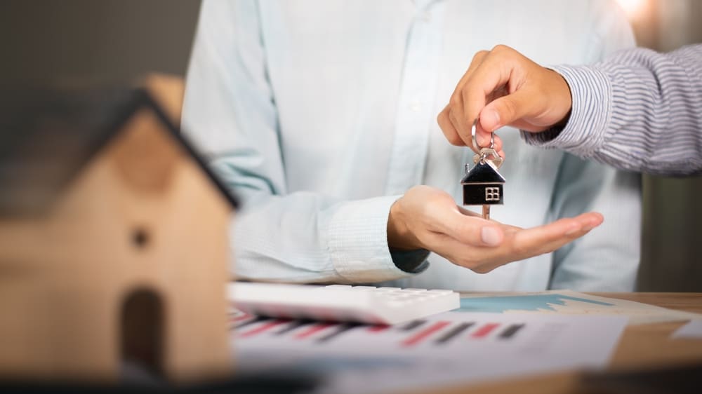 Hand passing a house-shaped key ring beside a model house